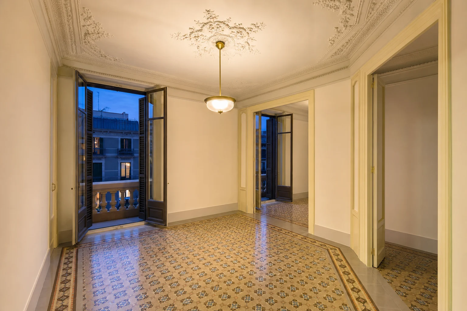 Estancia vacía de un piso del Eixample al anochecer, con las puertas de balcón abiertas al azul de la noche y la luz cálida de una lámpara de latón encendida